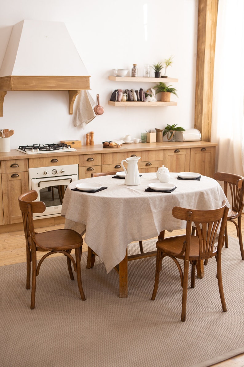 May include: A kitchen interior with wooden cabinets, a stove, and a white oven. A round table is set with a beige tablecloth, white plates, and black placemats. Four wooden chairs surround the table. A white pitcher and sugar bowl are on the table.