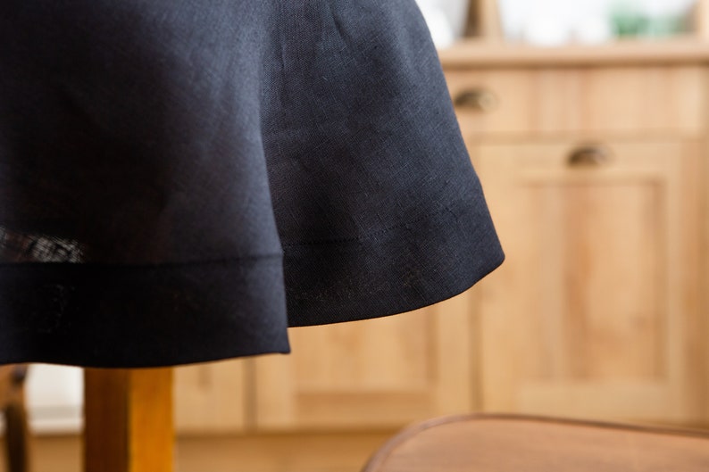 May include: Close-up of a dark navy blue linen tablecloth. The fabric drapes over a table, showcasing the texture and subtle sheen of the material. The hem is neatly stitched, adding a touch of elegance.