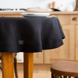 May include: A black round tablecloth draped over a wooden table, with a white plate on top. The tablecloth has a simple, elegant design. The background shows a kitchen with wooden cabinets and chairs.