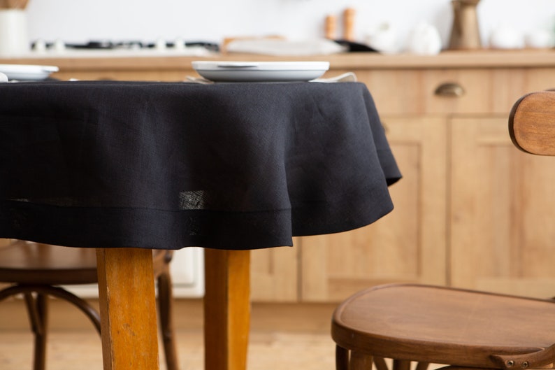 May include: A black round tablecloth draped over a wooden table, with a white plate on top. The tablecloth has a simple, elegant design. The background shows a kitchen with wooden cabinets and chairs.