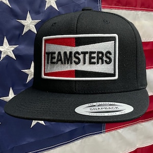 May include: Black and white baseball cap with a red, white, and black patch that says "TEAMSTERS" on it. The cap has a black snap closure and a white label that says "THE AUTHENTIC SNAPBACK YUPOONG SINCE 1974".