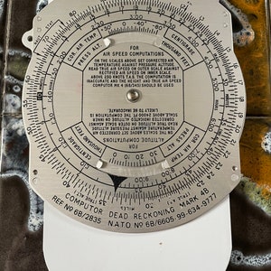 May include: A silver circular aviation flight computer with a black needle and markings. The computer is used for air speed computations and dead reckoning. The text "FOR AIR SPEED COMPUTATIONS" and "DEAD RECKONING" is printed on the face of the computer.