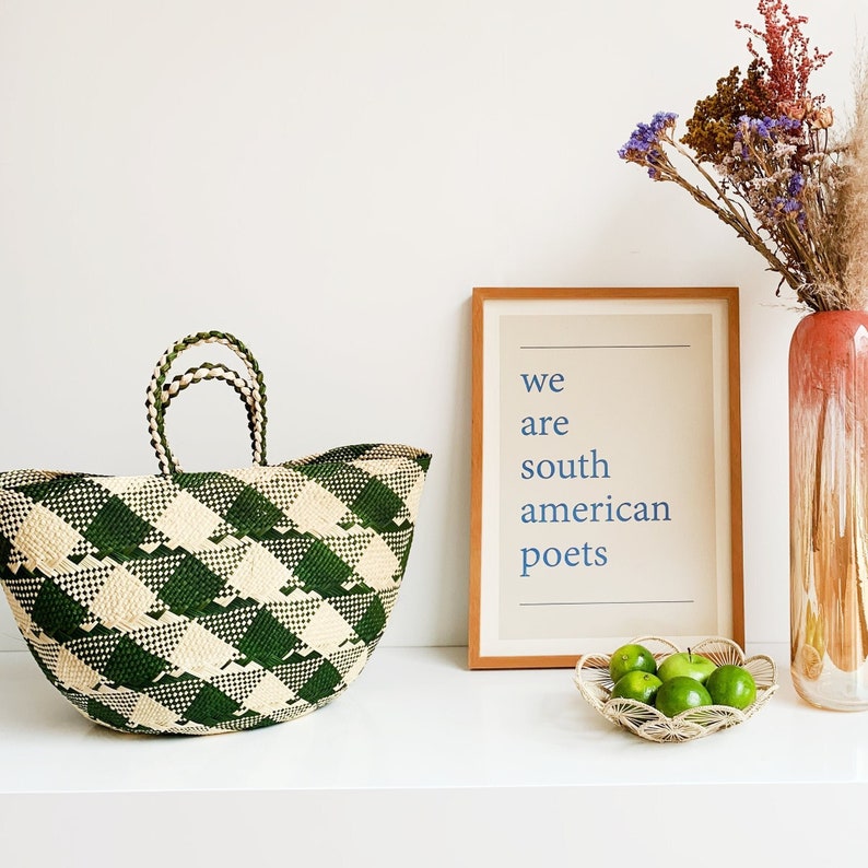 Puede incluir: Una bolsa de mano tejida a cuadros verdes y blancos con dos asas. La bolsa est&aacute; sobre una superficie blanca junto a una impresi&oacute;n enmarcada con el texto "we are south american poets".