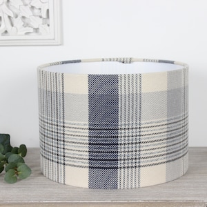 May include: A cream and blue plaid lampshade with a white lining. The lampshade is cylindrical and has a subtle texture.