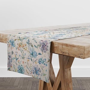 May include: A floral table runner with a watercolour design featuring blue, purple, and yellow flowers on a light beige background. The runner is laying on a wooden table.