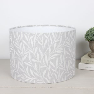 May include: A cylindrical lampshade featuring a light grey backdrop adorned with a repeating white leaf design. The interior of the lampshade is white. This lampshade is a home decor item.