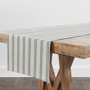 May include: A striped table runner in beige and grey-green, draped over a wooden table. The runner has vertical stripes, adding a rustic touch to the table setting. The table is made of light-coloured wood. The runner's length is approximately 150 cm.
