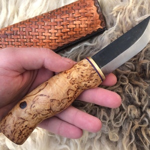 May include: A handcrafted knife with a light-colored, patterned wooden handle and a dark blade. The handle has a dark blue dot and is accented with gold and purple bands. A brown leather sheath is visible in the background.