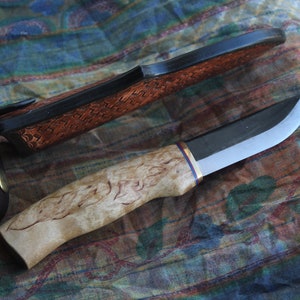 May include: A handcrafted knife with a light-colored, patterned wooden handle and a curved blade. The knife has a dark sheath with a woven leather design and a leather strap. The blade is silver and black.