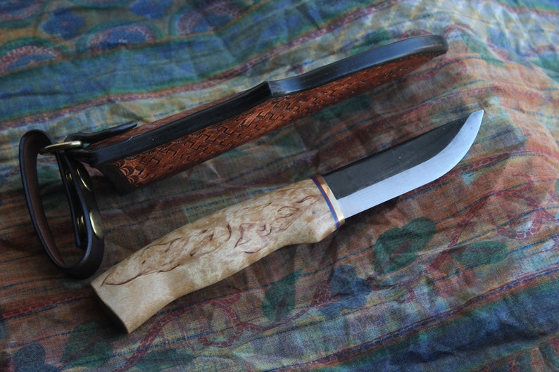 May include: A handcrafted knife with a light-colored, patterned wooden handle and a curved blade. The knife has a dark sheath with a woven leather design and a leather strap. The blade is silver and black.