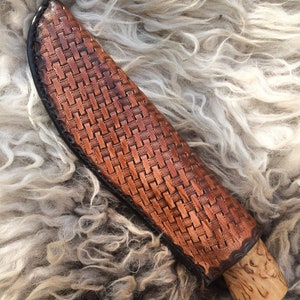 May include: A brown leather knife sheath with a woven basket pattern. The sheath has a black border and a wooden handle. The knife sheath is designed to protect the knife blade.