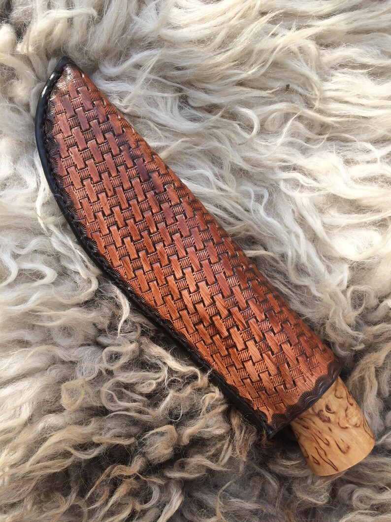 May include: A brown leather knife sheath with a woven basket pattern. The sheath has a black border and a wooden handle. The knife sheath is designed to protect the knife blade.