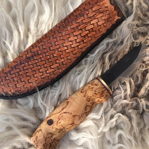 May include: A handcrafted knife with a dark blade and a light wood handle. The handle has a unique burl wood grain pattern. The knife is next to a brown leather sheath with a woven design. The knife and sheath are on a white fur surface.