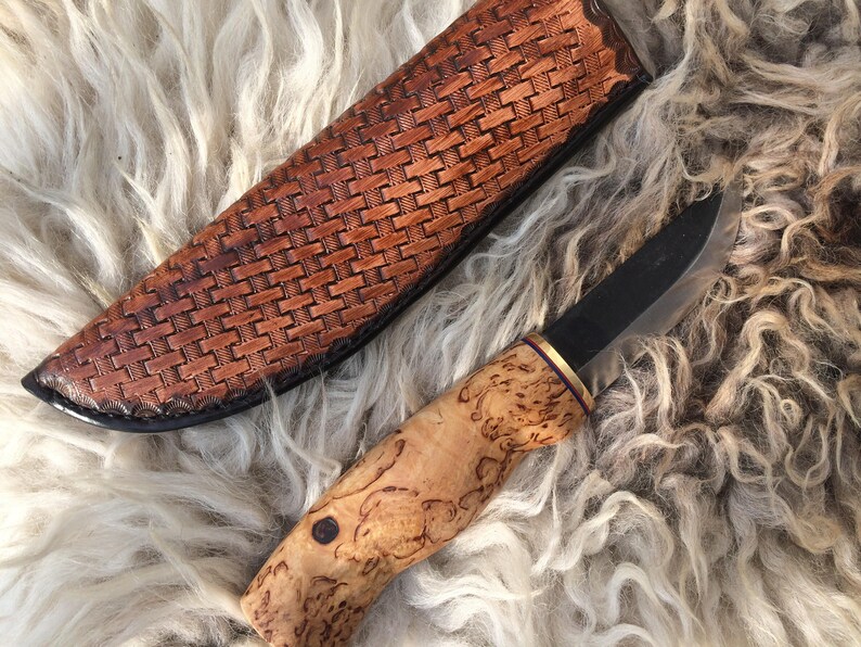 May include: A handcrafted knife with a dark blade and a light wood handle. The handle has a unique burl wood grain pattern. The knife is next to a brown leather sheath with a woven design. The knife and sheath are on a white fur surface.