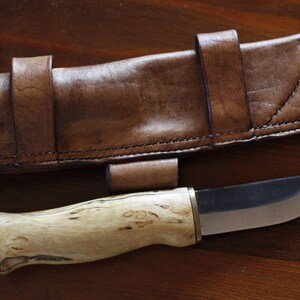 May include: A handcrafted knife with a light wood handle and a silver blade, accompanied by a brown leather sheath. The sheath has a belt loop and is stitched with dark thread. The knife and sheath are displayed on a wooden surface.
