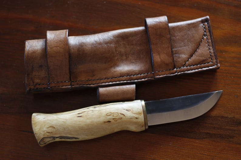 May include: A handcrafted knife with a light wood handle and a silver blade, accompanied by a brown leather sheath. The sheath has a belt loop and is stitched with dark thread. The knife and sheath are displayed on a wooden surface.