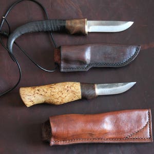 May include: Two handcrafted knives with leather sheaths and a leather cord. One knife has a dark, curved handle, the other a light-colored, textured handle. The blades are silver, and the sheaths are brown.