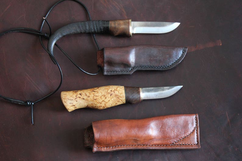 May include: Two handcrafted knives with leather sheaths and a leather cord. One knife has a dark, curved handle, the other a light-colored, textured handle. The blades are silver, and the sheaths are brown.