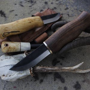 May include: A collection of handcrafted knives with wooden handles and dark blades. The knives are displayed on a textured surface, with a leather sheath and deer antlers adding to the rustic aesthetic. The handles vary in color, from light to dark brown.