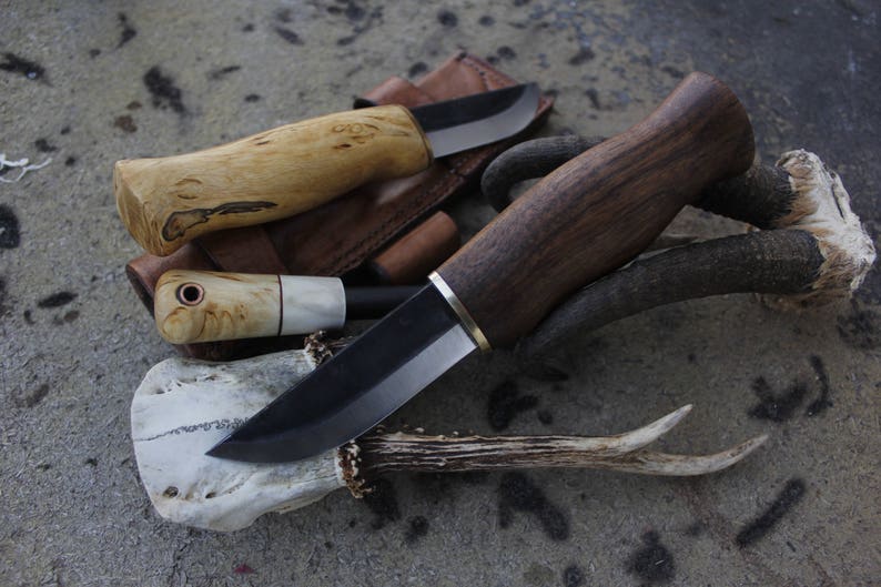 May include: A collection of handcrafted knives with wooden handles and dark blades. The knives are displayed on a textured surface, with a leather sheath and deer antlers adding to the rustic aesthetic. The handles vary in color, from light to dark brown.