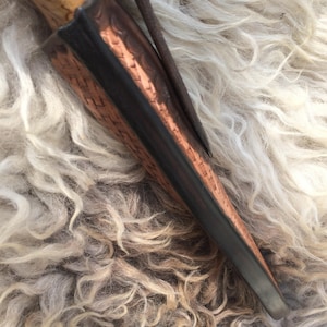 May include: A handcrafted knife with a light-colored wooden handle and a leather sheath. The sheath is brown with a dark border and a metal clip. The knife is partially inserted into the sheath, showcasing the blade's tip.
