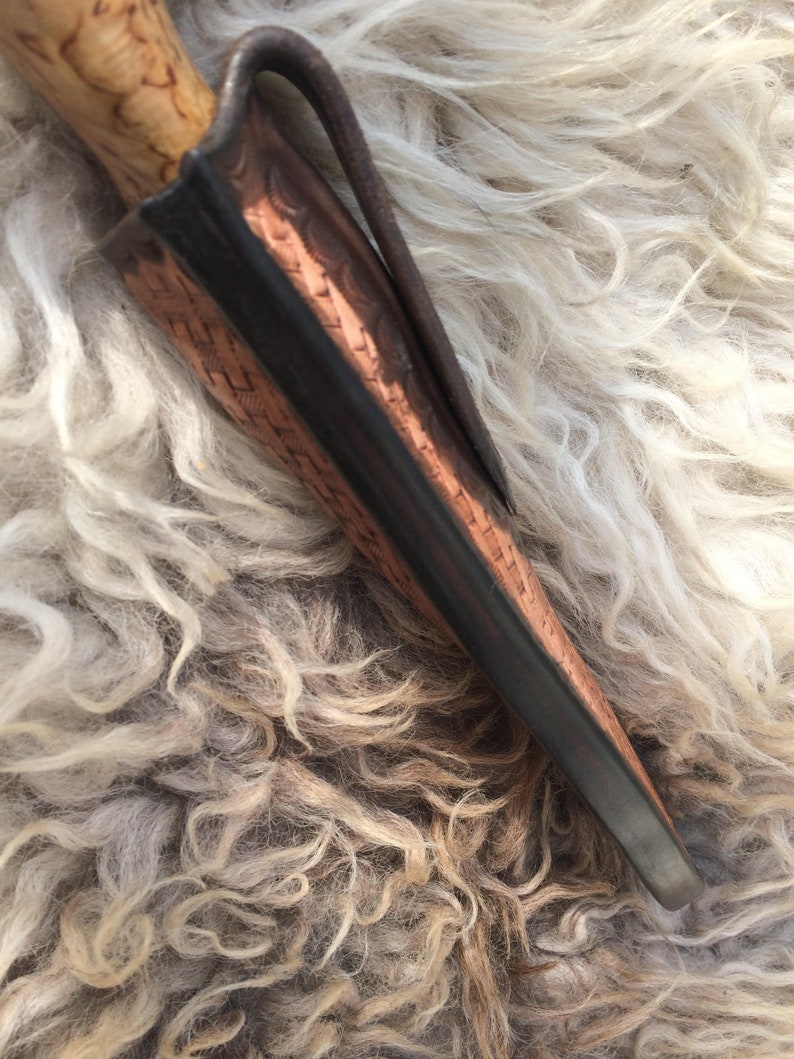 May include: A handcrafted knife with a light-colored wooden handle and a leather sheath. The sheath is brown with a dark border and a metal clip. The knife is partially inserted into the sheath, showcasing the blade's tip.