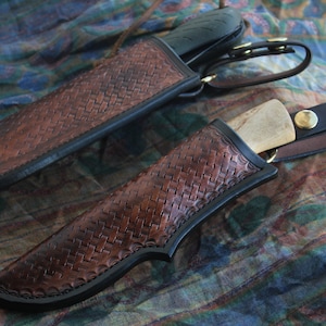 May include: Two handcrafted leather knife sheaths with a woven pattern in shades of brown and black. The sheaths have a black border and are secured with a leather strap and brass hardware. The knives have black handles and wooden blades.