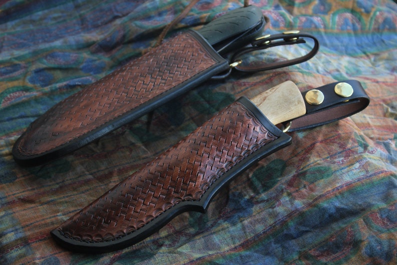 May include: Two handcrafted leather knife sheaths with a woven pattern in shades of brown and black. The sheaths have a black border and are secured with a leather strap and brass hardware. The knives have black handles and wooden blades.