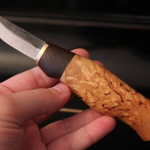 May include: A handcrafted knife with a polished steel blade and a handle made of light brown, patterned wood. The handle has a dark brown section near the blade and a brass-colored spacer. The knife is held in a hand.
