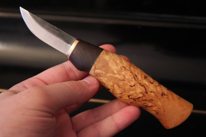 May include: A handcrafted knife with a polished steel blade and a handle made of light brown, patterned wood. The handle has a dark brown section near the blade and a brass-colored spacer. The knife is held in a hand.