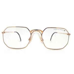 Puede incluir: Gafas de metal de tono dorado con lentes transparentes. La montura tiene una forma geométrica y las patillas están marcadas con "PORSCHE DESIGN".