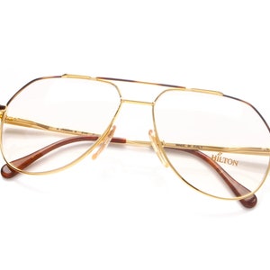 May include: Gold-tone aviator eyeglasses with brown tortoise shell temple arms. The glasses have a clear lens and say "HILTON" on the right arm. "MADE IN ITALY" is printed on the right arm.