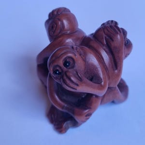 May include: A small, hand-carved wooden netsuke sculpture featuring three monkeys huddled together. One monkey covers its mouth with its hands, another covers its ears, and the third covers its eyes, all rendered in a smooth, polished brown wood.