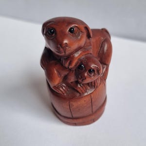 May include: A small, hand-carved brown wooden netsuke figurine depicting two monkeys. One larger monkey sits atop a wooden barrel, while a smaller monkey is positioned directly in front of it, resting its hands on the side of the barrel structure.