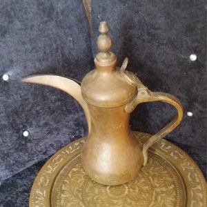 May include: A vintage brass coffee pot with a long spout and a curved handle, sitting on a matching brass tray. The tray has an intricate floral pattern. The pot and tray have a warm, aged patina.