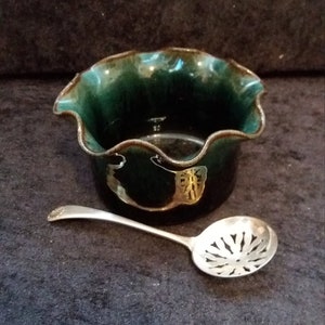 May include: A dark green ceramic bowl with a ruffled rim and a silver spoon. The bowl has a metallic sheen and a decorative design on the side. The spoon has a slotted design.