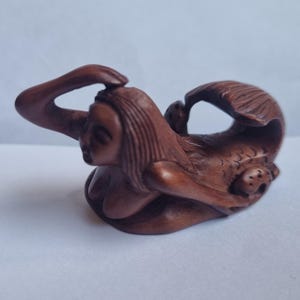 May include: A small, hand-carved brown wooden figurine depicting a reclining mermaid with long hair, one arm raised to the head, and a curved tail. The sculpture features detailed facial expressions and textured scales along the lower body.