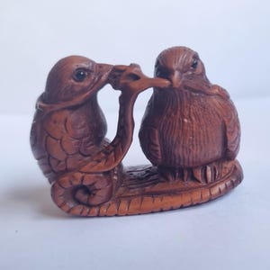 May include: A brown carved wooden figurine depicting two birds perched on a branch. One bird is positioned slightly behind the other, with their beaks touching. The carving features detailed textures on the feathers and the branch base.