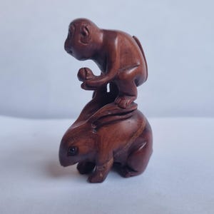 May include: A small, hand-carved wooden figurine featuring a monkey sitting atop a rabbit. The monkey is holding a small round object in its hands, while the rabbit is positioned below with its ears laid back. The entire piece has a smooth, dark brown finish.