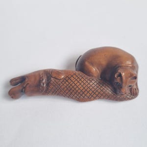 Op de afbeelding: Een bruin gesneden houten netsuke-beeldje dat een kat voorstelt die zijn kop op een grote vis laat rusten. De vis heeft een gedetailleerd kruislings schubbenpatroon langs het lichaam, terwijl de kat een gladde vacht en zichtbare gelaatstrekken heeft.