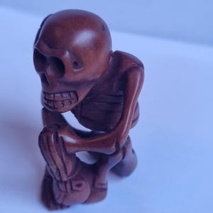 May include: A small, hand-carved brown wooden figurine depicting a skeleton in a kneeling position. The skeleton has a smooth, polished finish, visible rib cage details, and is positioned with its hands resting on its knees in a seated posture.