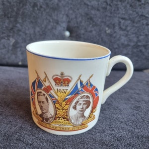 Peut inclure: Une tasse en céramique blanche avec un bord et une anse bleus. La tasse présente des portraits d'un homme et d'une femme, des drapeaux, une couronne et le texte "1937 Coronation Long May They Reign."