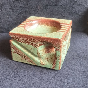 May include: A square ceramic ashtray with a light green base and reddish-brown accents. The ashtray has a circular indentation and a carved design on the side. The ashtray is sitting on a dark grey surface.