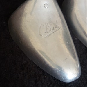 May include: Two silver-colored shoe trees with a metal rod and a curved, rounded toe shape. The word "Club" is engraved on the surface of the shoe tree. The background is a dark, textured fabric.