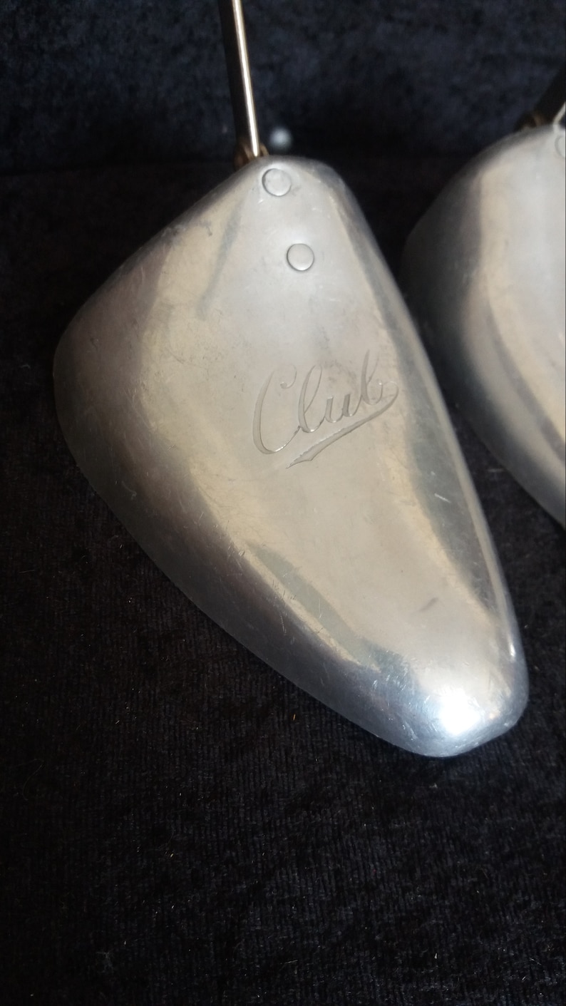May include: Two silver-colored shoe trees with a metal rod and a curved, rounded toe shape. The word "Club" is engraved on the surface of the shoe tree. The background is a dark, textured fabric.