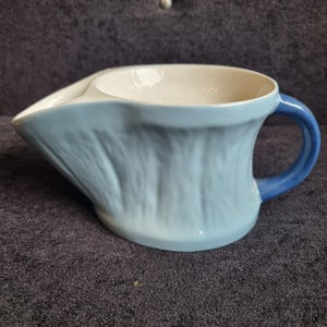 Puede incluir: Una salsera de cerámica azul claro con un asa azul. El interior es blanco. El exterior tiene un diseño texturizado y con volantes. La salsera está sobre una superficie gris oscuro.