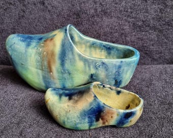 Pode incluir: Dois tamancos de cerâmica, um grande e outro pequeno, em tons de azul, verde e castanho. Os tamancos têm um acabamento vidrado e são provavelmente itens decorativos. O tamanco maior tem cerca de 20 cm de comprimento.