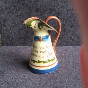 Puede incluir: Jarra de cerámica con asa y boquilla marrones, cuerpo blanco con detalles pintados en azul, verde y naranja. El texto "Home the spot on earth supremely blest" está escrito en la parte delantera.