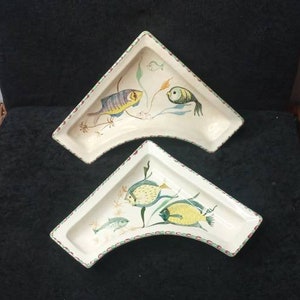 May include: Two decorative, triangular ceramic dishes with a fish motif. The dishes are off-white with colorful fish illustrations in yellow, blue, and green. The edges are trimmed with a red and green pattern. The dishes are placed on a dark surface.