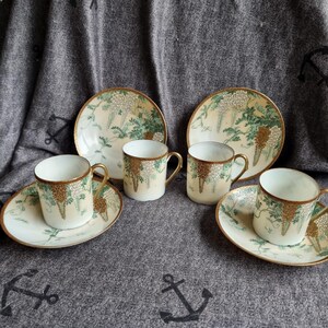 Puede incluir: Un juego de té vintage con tazas, platillos y platos. Las piezas color crema están adornadas con un diseño floral en verde y dorado, con bordes dorados. El conjunto se presenta sobre una tela gris decorada con motivos de anclas.